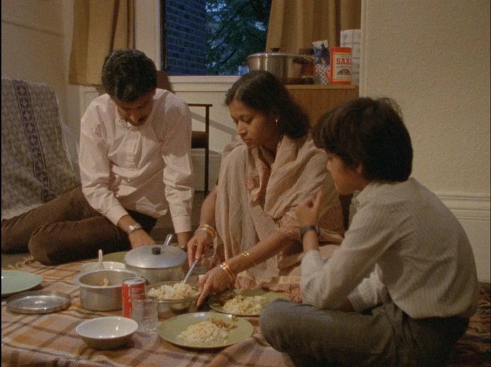A family of three seated on the floor, sharing a meal, with various dishes and a soda can on a tablecloth in a cozy room.