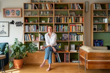Woman in white shirt and jeans sitting on a bookshelf ledge, holding a mug, surrounded by books and plants in a cozy living room.