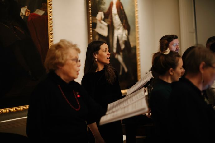 A choir sings enthusiastically in a gallery with framed portraits in the background.