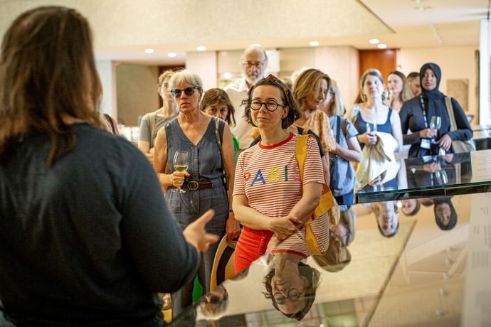 A diverse group of people attentively listens to a speaker in a well-lit indoor setting, with reflective surfaces capturing their images.
