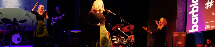 Singer performing on stage with a band, vibrant lighting, and a large screen displaying "barbican" in the background.