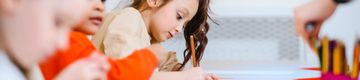 Young girl focused on drawing with colored pencils, surrounded by other children in a classroom setting.
