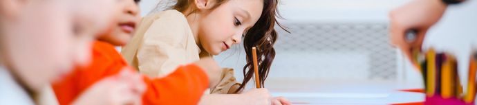 Young girl focused on drawing with colored pencils, surrounded by other children in a classroom setting.