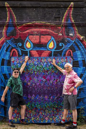 Two individuals in colourful shirts pose in front of a vibrant mural featuring a horned creature and a floral-patterned tapestry.