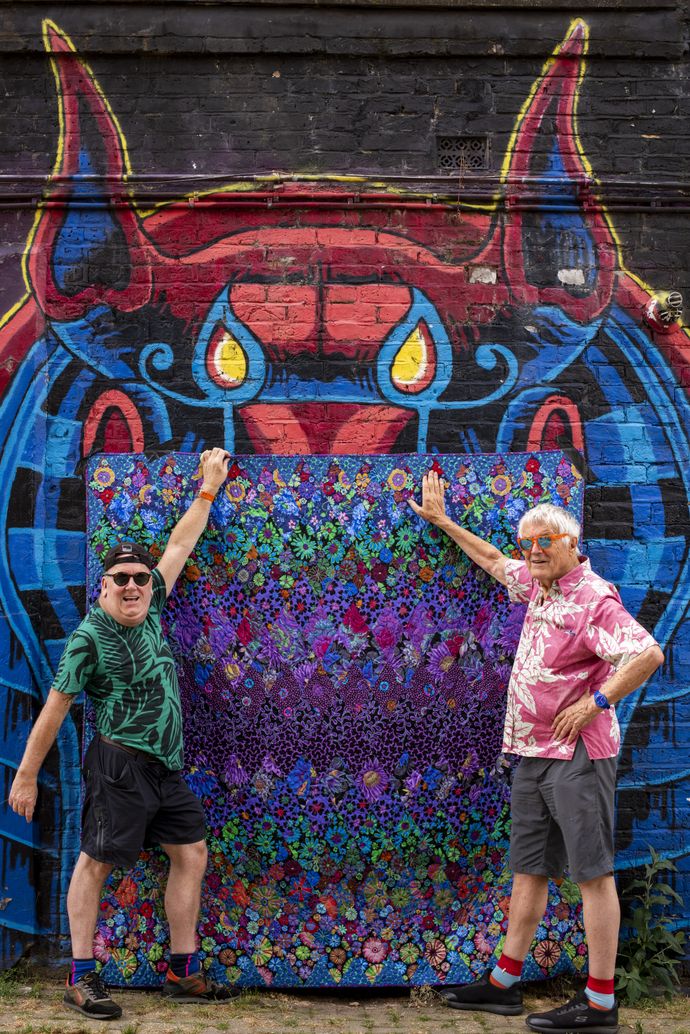 Two individuals in colourful shirts pose in front of a vibrant mural featuring a horned creature and a floral-patterned tapestry.