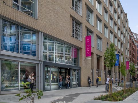 Modern brick building with large windows and "City Lit" signage, people entering and exiting, trees lining the sidewalk.