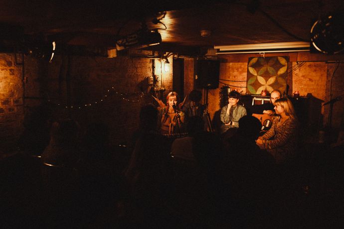 Intimate live music performance in a dimly lit, rustic venue with four musicians, surrounded by a cozy audience.