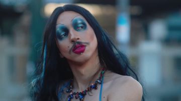 A person with long black hair, bold blue eyeshadow, dramatic makeup, and a striking necklace, looking sideways outdoors.