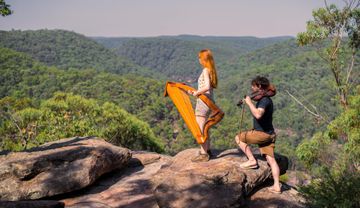 Two musicians play harp and violin on a rocky cliff, surrounded by lush green hills under a clear sky.