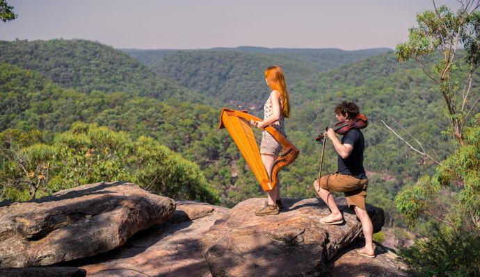 Two musicians play harp and violin on a rocky cliff, surrounded by lush green hills under a clear sky.