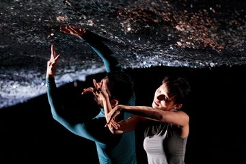 Two dancers in dynamic poses reach towards a textured, dark ceiling. The scene is dramatic and artistic, with contrasting lighting.