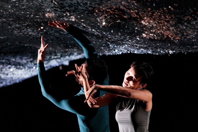 Two dancers in dynamic poses reach towards a textured, dark ceiling. The scene is dramatic and artistic, with contrasting lighting.