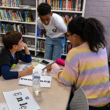 Three children in a library work together on a project at a table with bookshelves in the background.