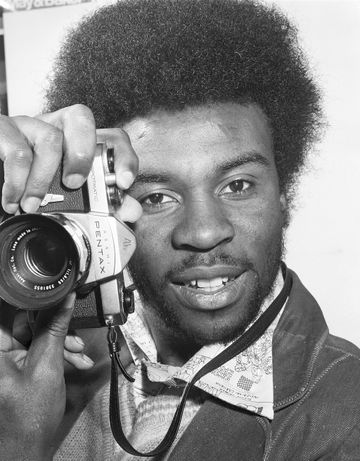 Person holding a Pentax camera, looking directly at the viewer, wearing a patterned shirt and jacket.