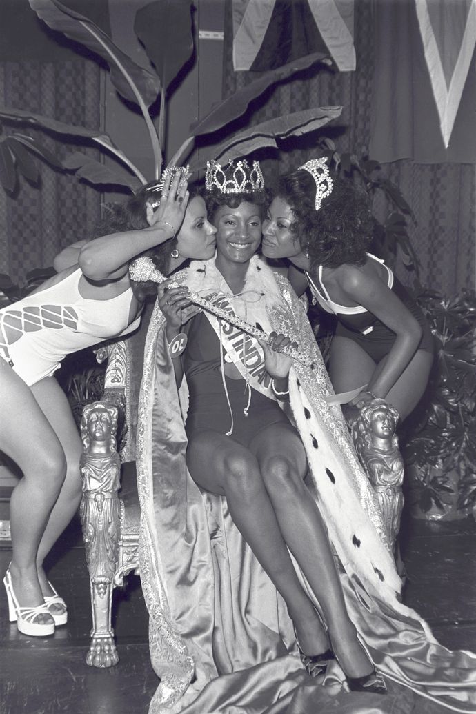 Three women in swimsuits and crowns celebrate on stage; the seated woman wears a cape and crown, smiling as the others kiss her cheeks.