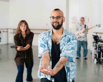 A person in a blue shirt gestures with hands outstretched, while a woman in the background smiles and a man with a guitar stands behind them.