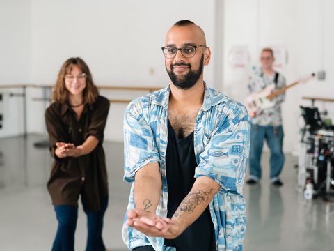 A person in a blue shirt gestures with hands outstretched, while a woman in the background smiles and a man with a guitar stands behind them.