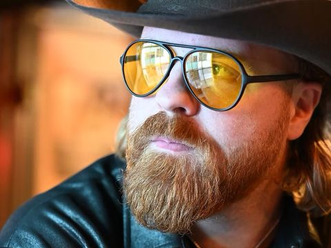 Person with a red beard wearing a wide-brimmed hat, yellow-tinted sunglasses and a dark shirt indoors.
