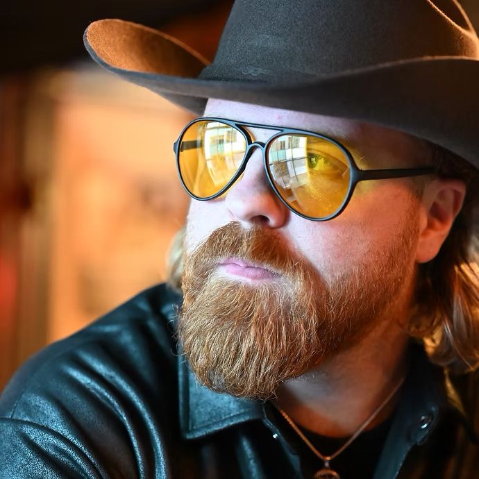 Person with a red beard wearing a wide-brimmed hat, yellow-tinted sunglasses and a dark shirt indoors.