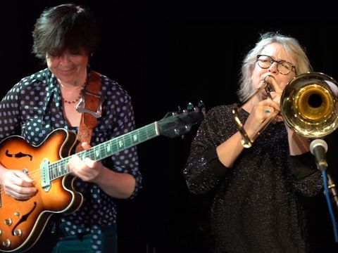 Two musicians performing on stage: one playing an electric guitar, and the other playing a trombone, both deeply focused on their music.