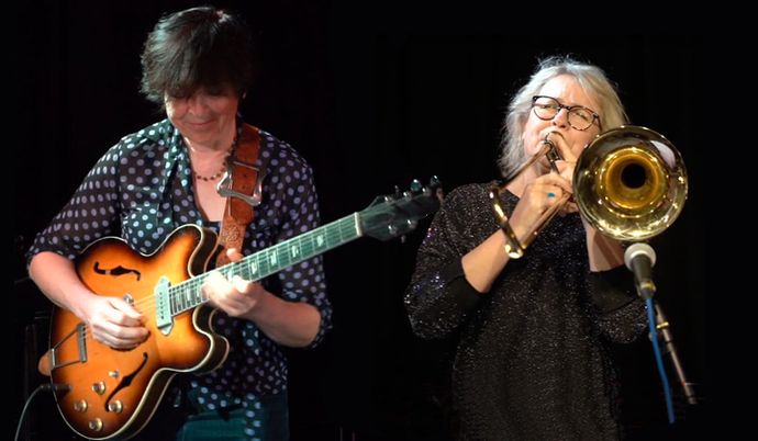 Two musicians performing on stage: one playing an electric guitar, and the other playing a trombone, both deeply focused on their music.
