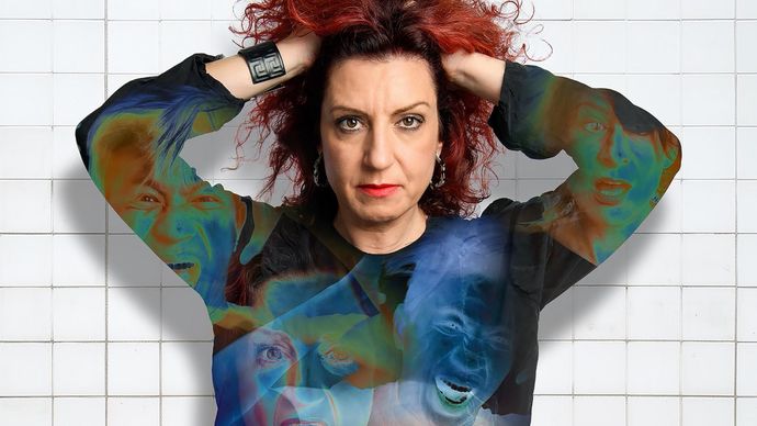 Person with curly red hair, hands on head, wearing a shirt with various expressive faces, standing against a white tiled background.