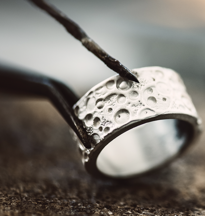 Close-up of a silver ring with a textured, crater-like surface, being crafted or examined with a pointed tool on a rustic background.