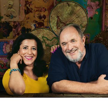 One person with pink nails and yellow top smiles to camera next to man with white beard and navy top sits smiling with arms folded