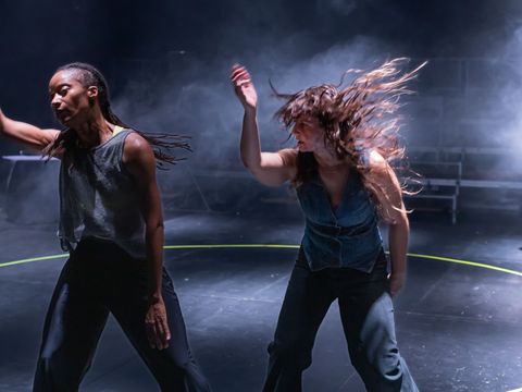 Two people stand in black room wearing sleeveless tops and black trousers - they are both in swing of movement with right arms reaching up and long hair flicking in the air