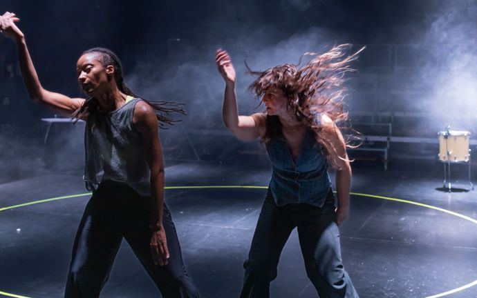 Two people stand in black room wearing sleeveless tops and black trousers - they are both in swing of movement with right arms reaching up and long hair flicking in the air
