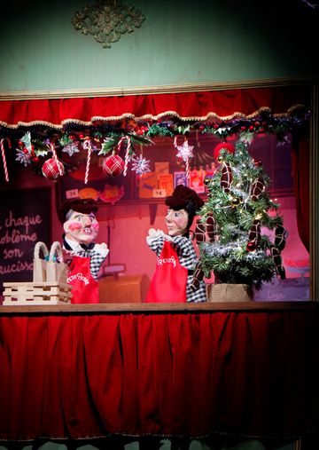 Two puppets in red aprons perform on a stage decorated with a small Christmas tree, candy canes, and festive ornaments.