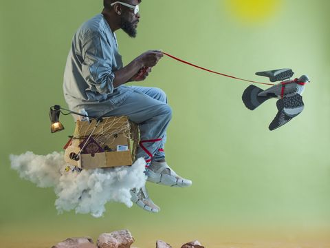 A man sitting on a floating cloud with a cardboard box, wearing a red hat and sunglasses, holds a leash attached to a flying robotic bird.