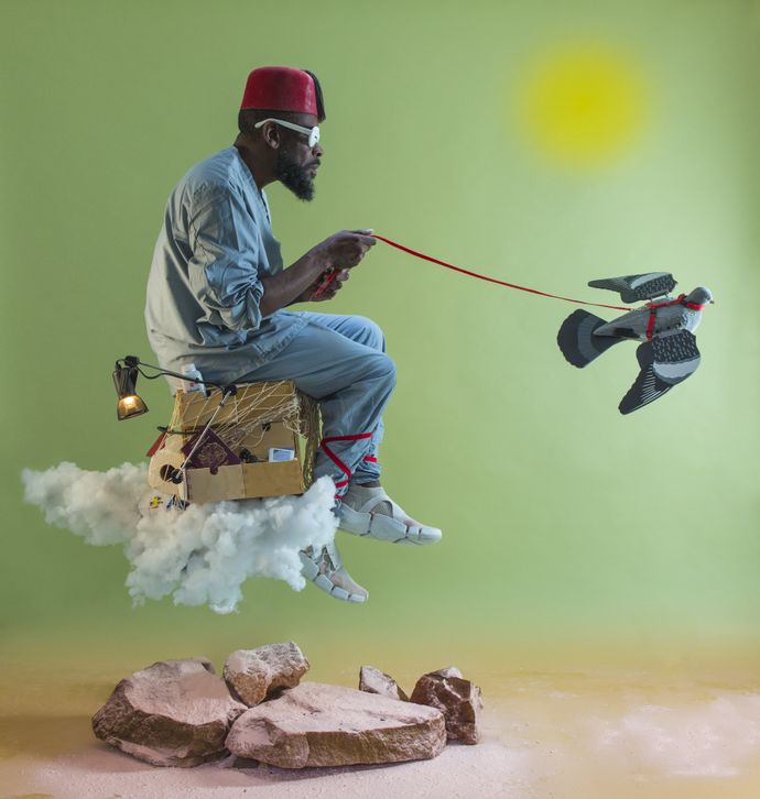 A man sitting on a floating cloud with a cardboard box, wearing a red hat and sunglasses, holds a leash attached to a flying robotic bird.
