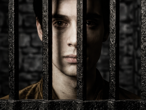 A person stands behind iron bars, face partially lit, with a stone wall in the background.