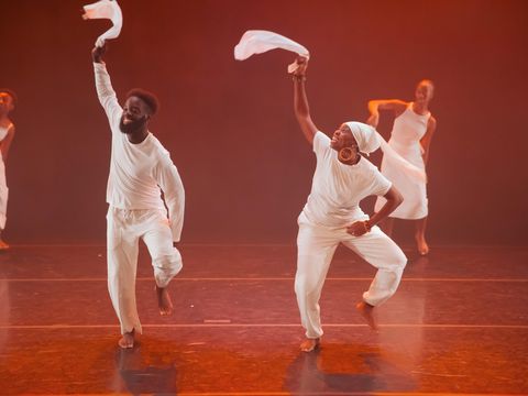 Room lit in reddish orange with 4 people wearing white tops and trousers lifting their hands in air holding white material, smiling in movement