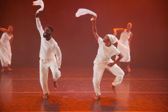 Room lit in reddish orange with 4 people wearing white tops and trousers lifting their hands in air holding white material, smiling in movement