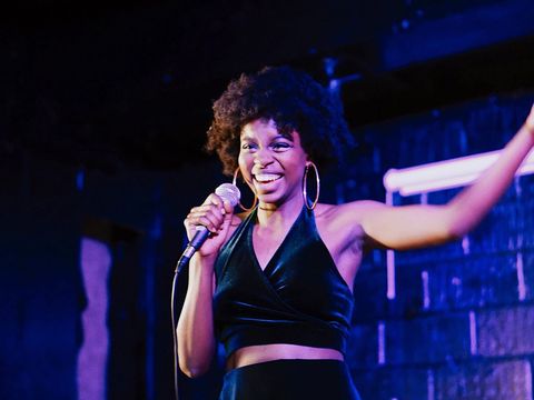 Person in a sleeveless black top holding a microphone and smiling on stage with one arm raised