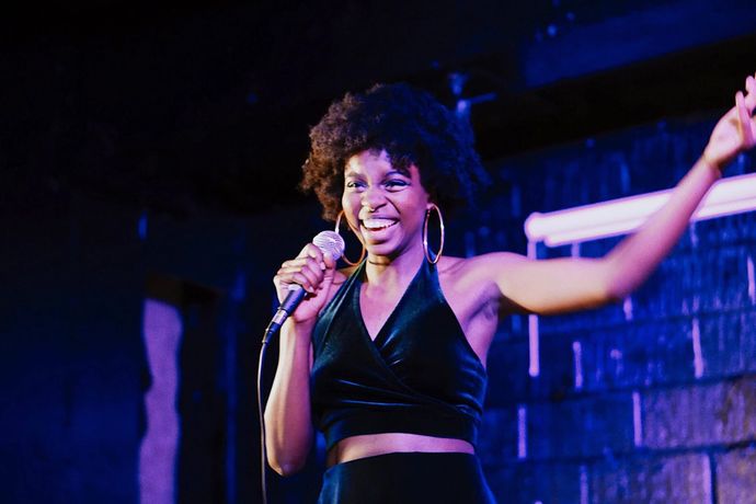 Person in a sleeveless black top holding a microphone and smiling on stage with one arm raised