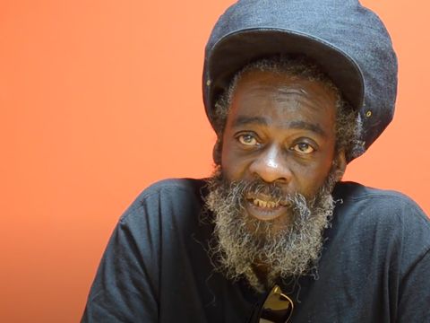 Person with a beard wearing a large denim hat and dark shirt sits against an orange background, looking towards the camera.