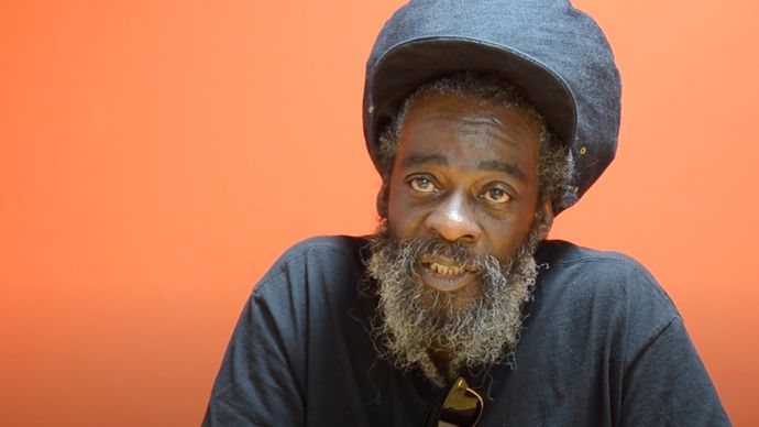 Person with a beard wearing a large denim hat and dark shirt sits against an orange background, looking towards the camera.