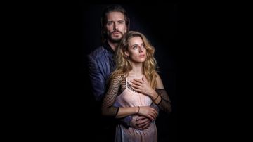 Two people posed together against dark background, one standing behind, arms wrapped around front.