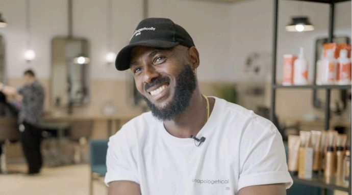 Smiling individual with a beard and cap sits in a salon, wearing a white t-shirt. Shelves with hair products are in the background.