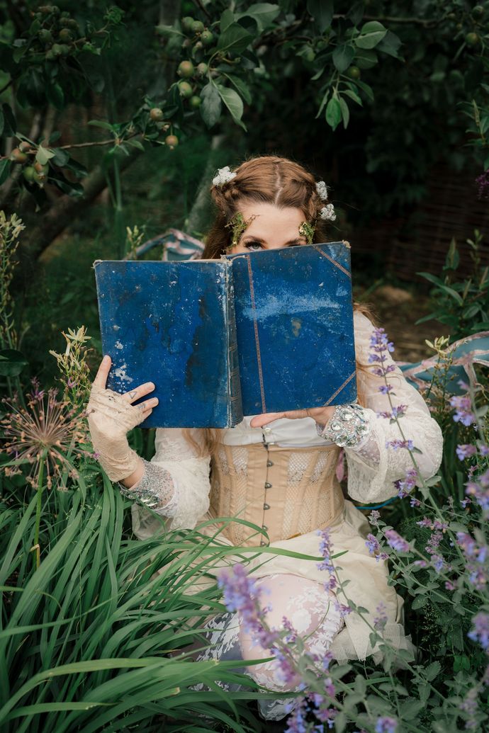 Person in vintage attire holding a large blue book, partially hidden by foliage and surrounded by blooming purple flowers.