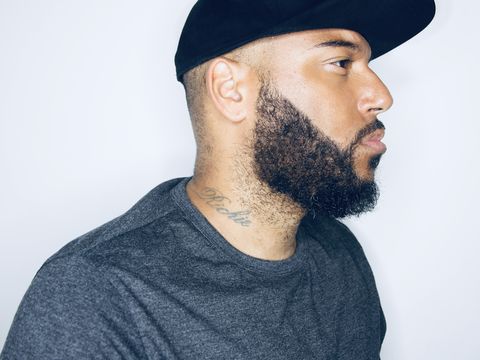 Person with a trimmed beard and neck tattoo wearing a black cap and grey shirt, shown in profile against a plain light background.