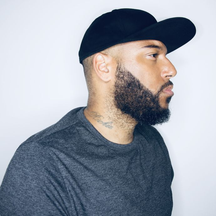 Person with a trimmed beard and neck tattoo wearing a black cap and grey shirt, shown in profile against a plain light background.