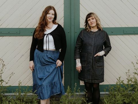 Two women stand in front of a green and white barn door, wearing denim outfits with plants in the foreground.