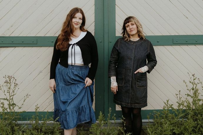 Two women stand in front of a green and white barn door, wearing denim outfits with plants in the foreground.