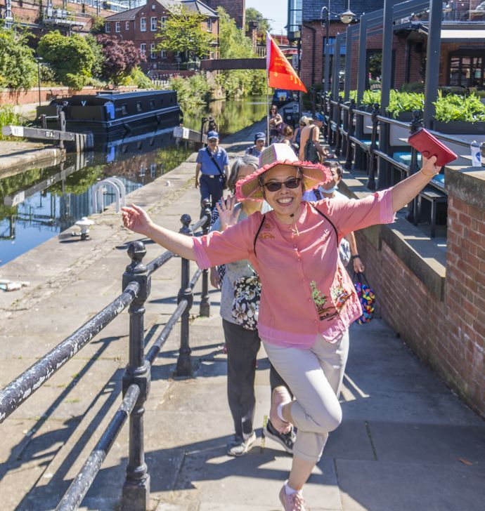 Smiling person in pink blouse and straw hat posing energetically on a canal towpath, group behind her.