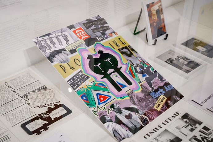 A colourful collage poster with abstract figures and cut-out images, surrounded by documents and black-and-white photographs displayed in a glass case.