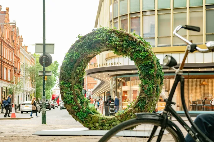 A circular living sculpture covered in greenery and plants displayed on a busy urban street near shops and pedestrians.